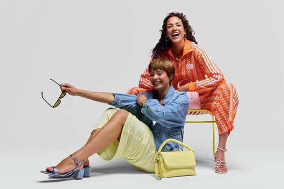 Zwei Frauen in bunten Frühlingsoutfits posieren mit Sonnenbrille, Handtasche und Sandalen vor hellem Hintergrund