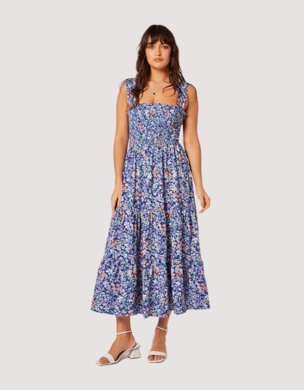 Frau in langem blauen Blumenkleid und Sandalen steht frontal und blickt ruhig in die Kamera