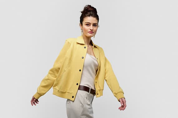 Frau trägt gelbe Jacke, weißes Top und beige Hose, posiert mit ausgestreckten Armen.