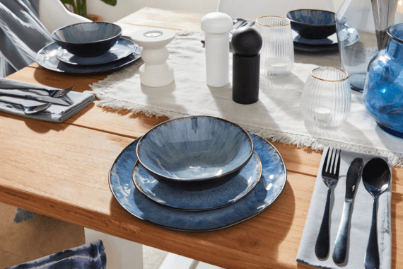 Gedeckter Holztisch mit blauem Teller-Set, Schalen, Besteck auf Stoffservietten sowie Salz- und Pfeffermühle